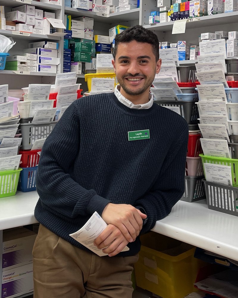 The founder of Izdyhar, a pharmacist, leaning against the counter of a dispensary lined with prescription baskets.